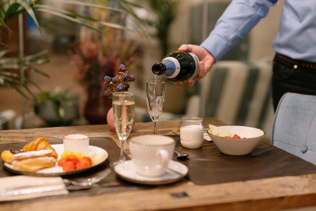 Een bediende schenkt bubbels in een glas bij een uitgebreid ontbijtbuffet met croissants, fruit en yoghurt